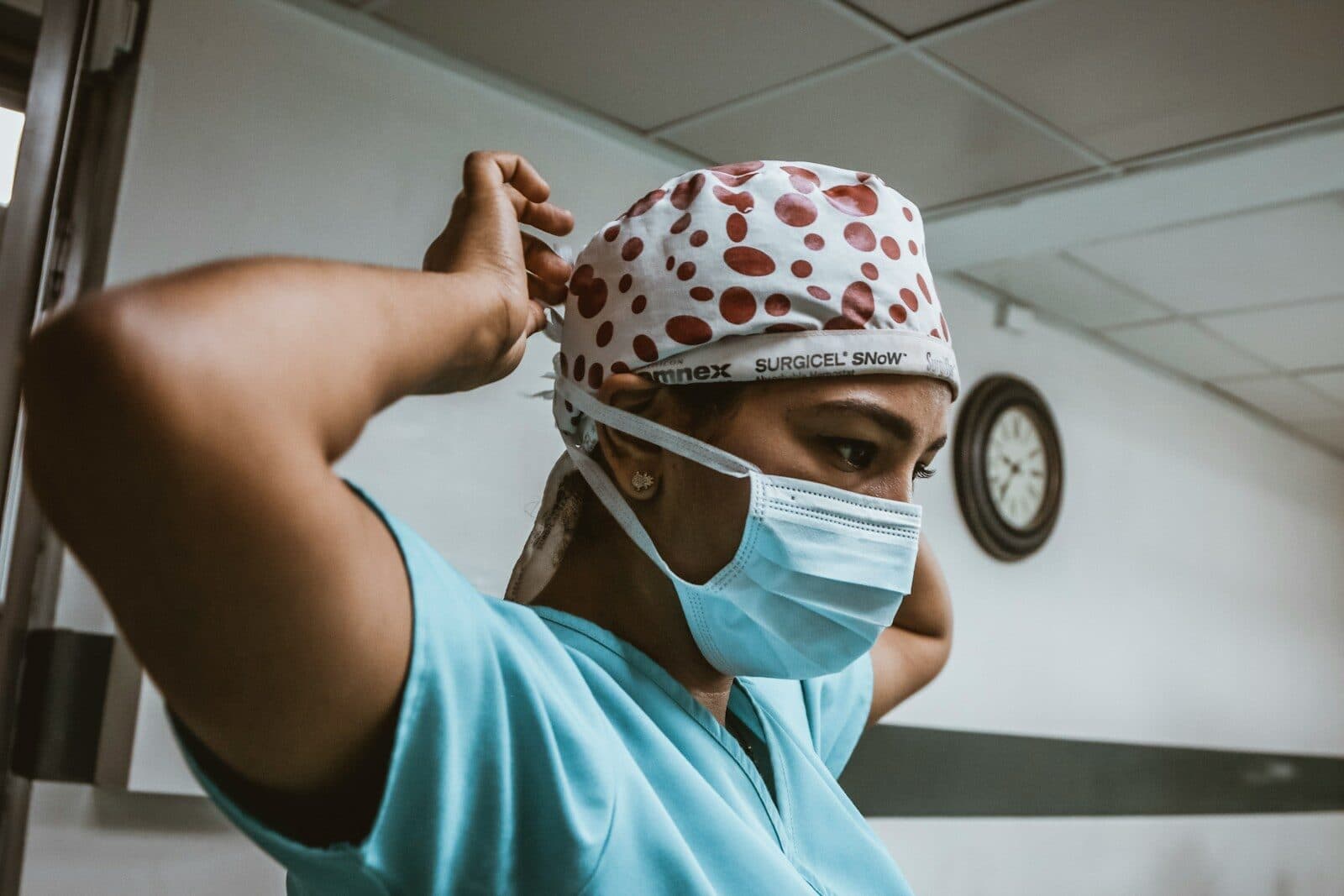 Trabajadora de la salud con ambo, cofia y barbijo se ata el barbijo en un pasillo, con un reloj de pared detrás.